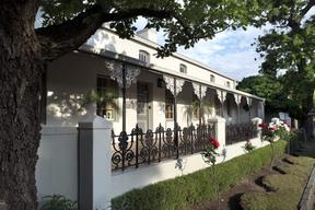 Front of white hotel building with decorative railings and grass in front. two trees to the forefront of the image.