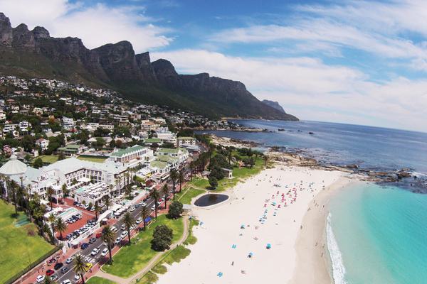 Aerial view of buildings with white sandy beach leading onto a turquoise sea, blue sky with some white clouds.