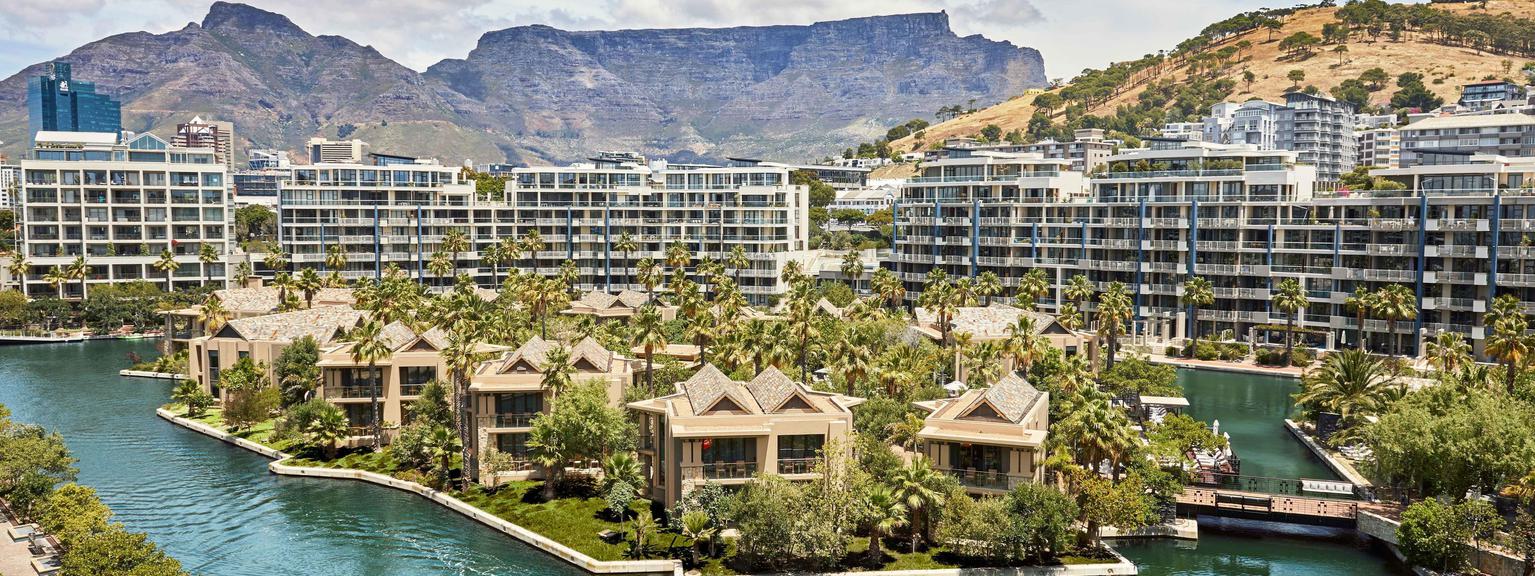 Aerial view of hotel, surrounded by blue water, with trees. Table Mountain is in the background.
